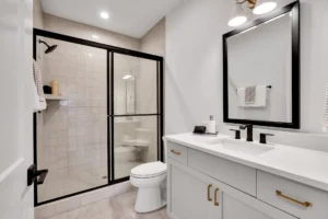 Modern white bathroom with black-framed glass shower, gold accents, and sleek minimalist design.