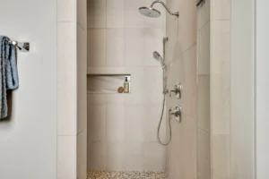 Modern beige tile walk-in shower with rainfall showerhead, pebble floor, and built-in niche.