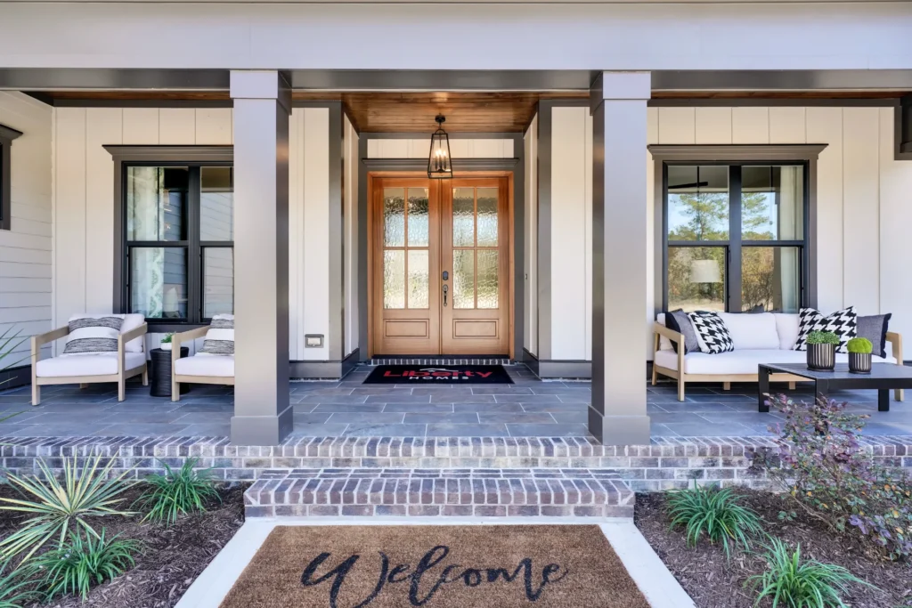 Inviting modern farmhouse porch with wooden double doors, lantern light, cozy seating, and greenery.