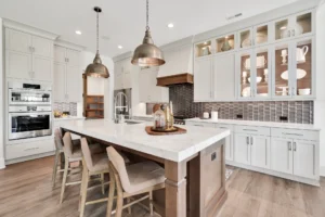 Modern white kitchen with marble island, pendant lights, and stainless steel appliances.