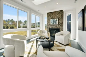 Bright modern living room with white armchairs, brick fireplace, and large sunlit windows.