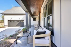 Modern front porch with wooden ceiling, stylish seating, and black-trimmed windows under blue skies.