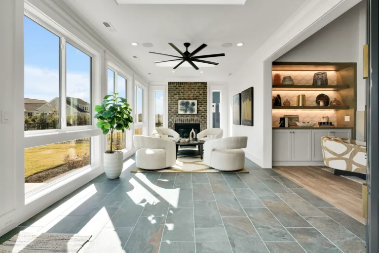 Bright modern sunroom with stone fireplace, white chairs, and large windows filled with natural light.