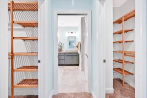 Bright master closet with light wood shelving leading to modern blue-gray bathroom.