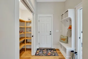Warm entryway mudroom with white bench, floral rug, and wooden floors leading to pantry shelves.