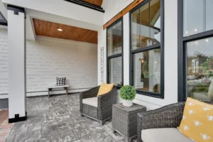 Modern farmhouse porch with wicker chairs, yellow pillows, stone flooring, and wood-paneled ceiling.