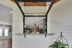 Bright modern kitchen with black metal pass-through window, white cabinets, and stone countertop.