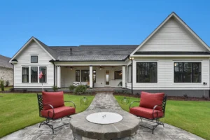 Modern farmhouse backyard with white siding, stone fire pit, and red cushioned patio chairs.