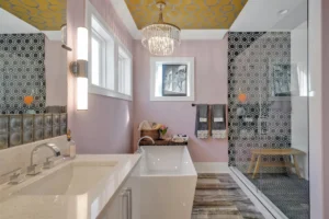 Modern pastel pink bathroom with gold ceiling, freestanding tub, chandelier, and elegant black‑white shower.