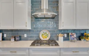 Modern white kitchen with blue tile backsplash, stainless cooktop, and gold hardware accents.
