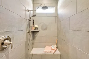 Modern beige tile walk-in shower with chrome rainfall head, built-in bench, and soft pink towels.