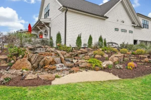 Modern farmhouse exterior with rock waterfall, landscaped garden, and lush lawn under blue sky.