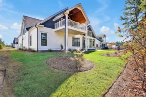Modern white farmhouse with dark trim, covered balcony, green lawn, and clear blue sky.
