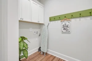 Modern laundry room with white cabinets, green rack, ironing board, and potted plant.
