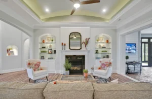 Bright elegant living room with fireplace, built-in shelves, light furniture, and large sunny windows.
