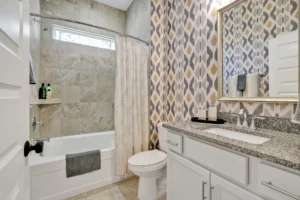 Modern gray tile bathroom with granite vanity, patterned wallpaper, and warm natural light.