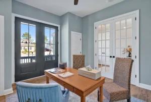 Coastal home office with light blue walls, wooden desk, French doors, and natural sunlight.