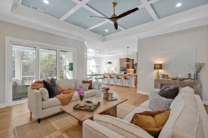 Modern open-concept living room with light blue coffered ceiling, beige sofas, and wood accents.
