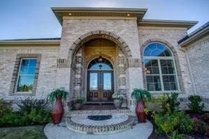 Elegant light brick home with arched wooden double doors and symmetrical landscaping.