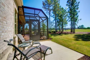 Modern brick home patio with lounge chairs and screened porch overlooking green lawn and pine trees.