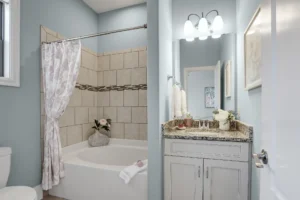 Light blue bathroom with granite vanity, tiled tub-shower, floral curtain, and soft natural lighting.