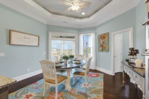 Coastal dining room with seafoam blue walls, wooden table, white chairs, and colorful floral rug.
