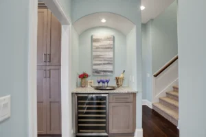 Elegant hallway home bar with arched niche, blue-gray walls, light wood cabinets, and wine fridge.