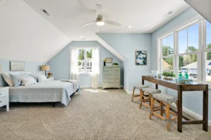Bright coastal bedroom with soft blue walls, natural light, cozy bed, and wooden desk.