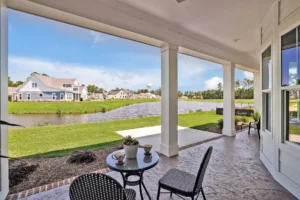 Covered porch with bistro set overlooking pond, green lawns, and suburban homes under clear sky.
