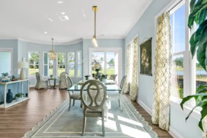 Elegant coastal dining room with glass table, blue walls, and sunlight through large windows.