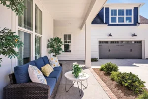 Modern coastal home exterior with blue accents, wicker seating, potted plants, and gray garage doors.