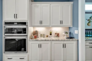 Modern white kitchen with blue walls, marble counters, double ovens, and warm under-cabinet lighting.