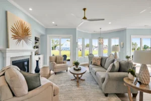 Bright coastal living room with light blue walls, beige furniture, and sunlit white fireplace.