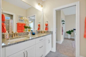 Bright modern bathroom with double vanity, gold faucets, white cabinets, and orange towel accents.