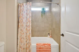 Modern bathroom with tan tiles, floral curtain, hanging plant, and bright orange towel.