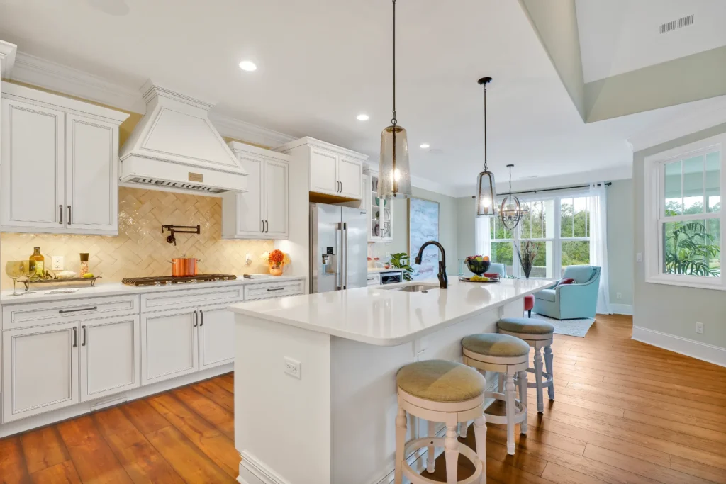 Modern white kitchen with quartz island, hardwood floors, pendant lights, and stainless steel appliances.