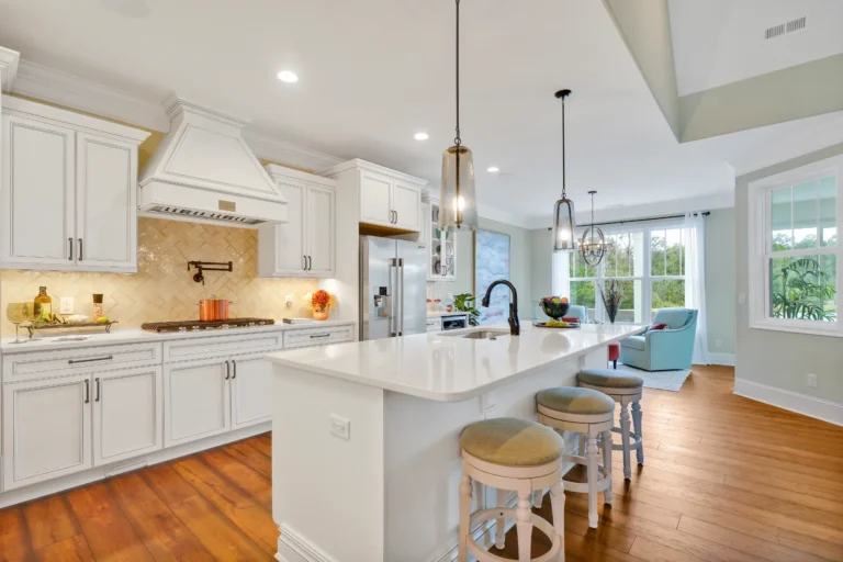 Modern white kitchen with quartz island, hardwood floors, pendant lights, and stainless steel appliances.