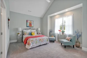 Bright gray bedroom with floral bedding, blue chairs, and sunlight filtering through white curtains.