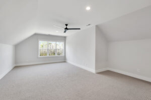 Bright minimalist attic room with white walls, beige carpet, large window, and modern black ceiling fan.