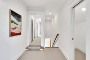 Modern minimalist hallway with white walls, beige carpet, abstract artwork, and bright natural lighting.
