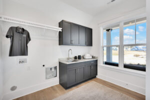 Bright modern laundry room with gray cabinets, white walls, and natural light from large window.
