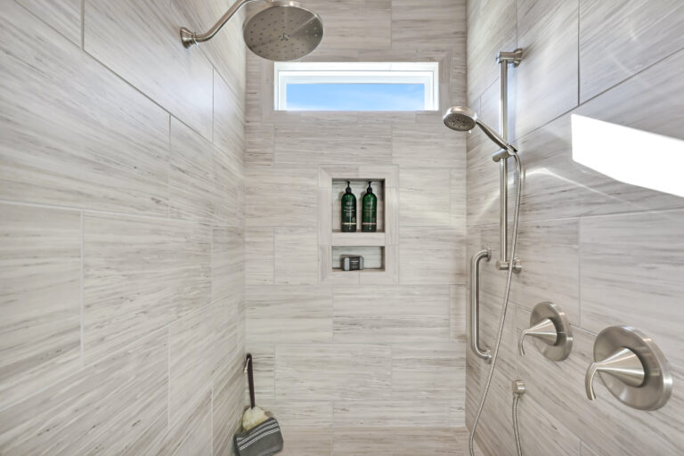 Modern gray tile walk-in shower with rainfall showerhead, handheld sprayer, and built-in shelf.