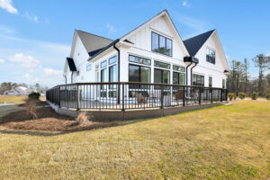 Modern white farmhouse with black windows, raised deck, and gabled roof under blue sky.