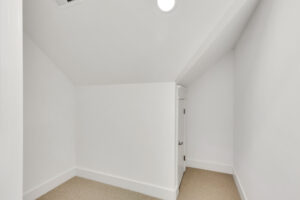 Bright white under-stair nook with slanted ceiling, beige carpet, and small storage door.