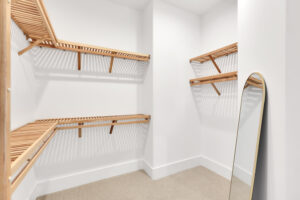 Bright minimalist walk-in closet with light wood shelving, white walls, and a gold-framed mirror.