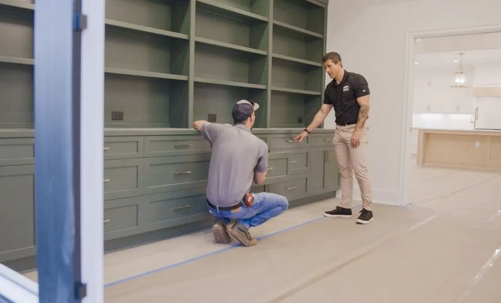 Two workers inspect custom olive green cabinetry and shelving built into a wall.
