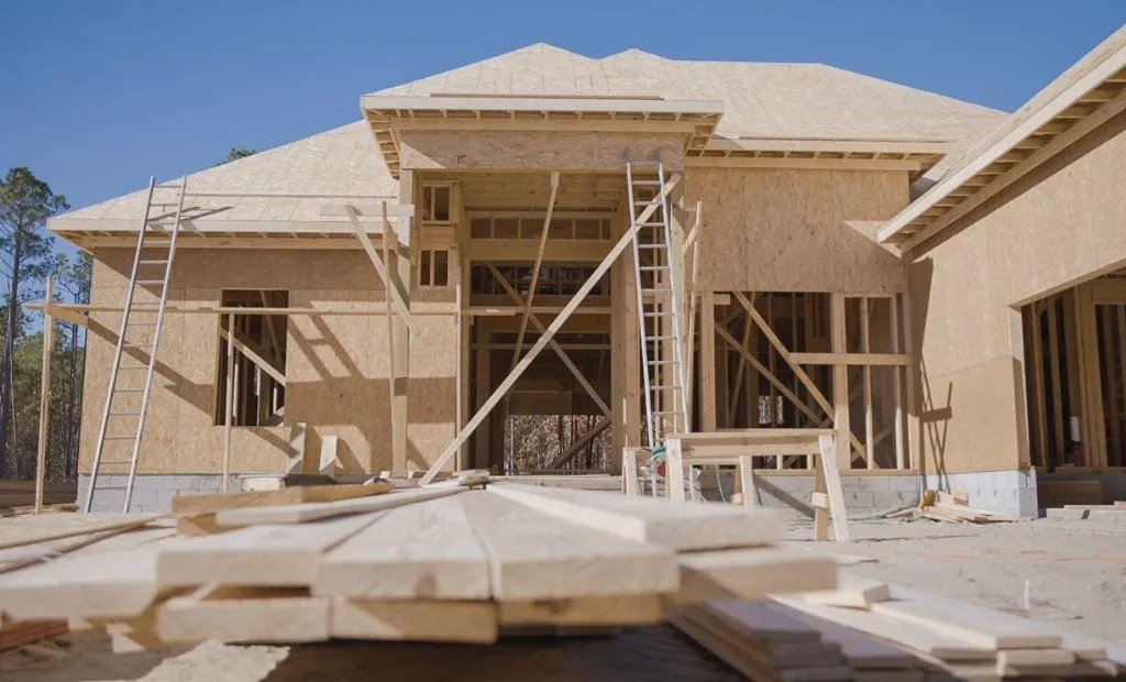 Exterior view of a stick-built home under construction with exposed wooden framing, sheathing, and stacked lumber in the foreground.